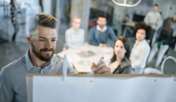 Young businessman writing ideas on whiteboard during a meeting with his colleagues. Young entrepreneur drawing business plan on whiteboard to his colleagues in the office. The view is through glass.